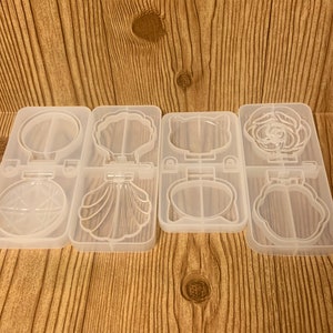 May include: Four clear silicone molds for resin casting. Each mold has two cavities. The top row features a circle, a seashell, a cat face, and a rose. The bottom row features a starburst, a seashell, a cat face, and a rose.
