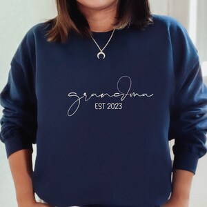 May include: A navy blue sweatshirt with the text "grandma est 2023" in white script.