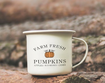 Puede incluir: Taza de esmalte blanco con un gráfico negro y naranja que dice "Farm Fresh Pumpkins Apples Hayrides Cider". La taza está sentada sobre una pila de troncos de árboles.