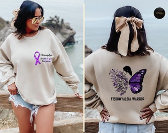 Op de afbeelding: Beige sweatshirt met een paars lint en de tekst "Fibromyalgie, Onzichtbaar voor jou, Echt voor mij". De achterkant van de sweatshirt heeft een paars vlinderontwerp met de woorden "Fibromyalgie Warrior".