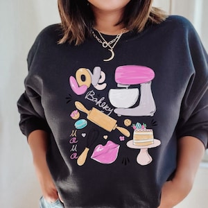 May include: Black sweatshirt with a graphic design featuring a pink and gray stand mixer, a rolling pin, a whisk, a cake, a heart, and the words "Love Bakery Mama".