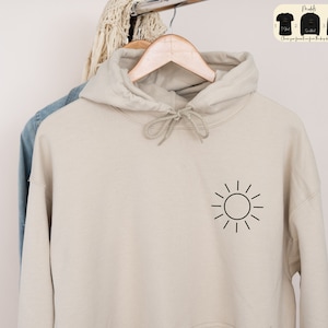 May include: A beige hoodie with a black sun graphic on the front. The hoodie has a drawstring hood and a kangaroo pocket.