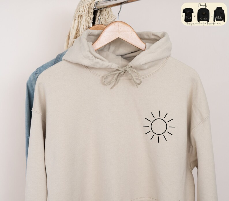 May include: A beige hoodie with a black sun graphic on the front. The hoodie has a drawstring hood and a kangaroo pocket.