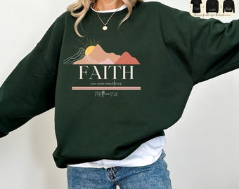 Puede incluir: Una sudadera verde oscuro con un gráfico de una cordillera y un sol con el texto "FAITH can move mountains Matthew 17:20".