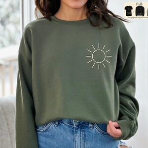 May include: A green sweatshirt with a white sun graphic on the front. The sweatshirt is a relaxed fit and has long sleeves.