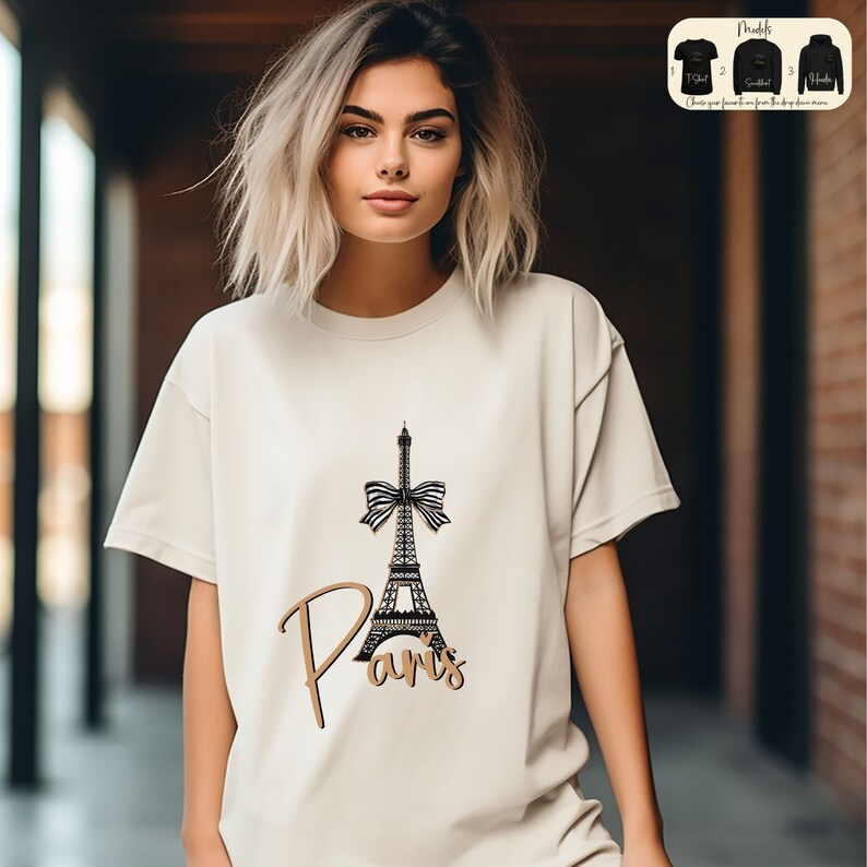 May include: A beige t-shirt with a black and white illustration of the Eiffel Tower with a black bow on top. The text "Paris" is written in gold script below the tower.