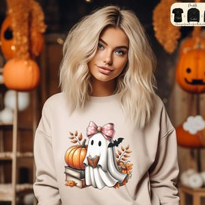 Puede incluir: Una sudadera beige con un gráfico de un fantasma con un lazo rosa leyendo un libro. El fantasma está sentado en una pila de libros con una calabaza y hojas de otoño alrededor.