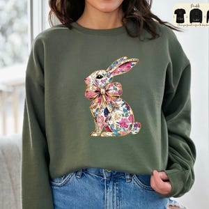Può includere: Una felpa verde con un coniglio con un motivo floreale che indossa un fiocco rosa. Il coniglio è seduto sulle zampe posteriori e ha le orecchie dritte.