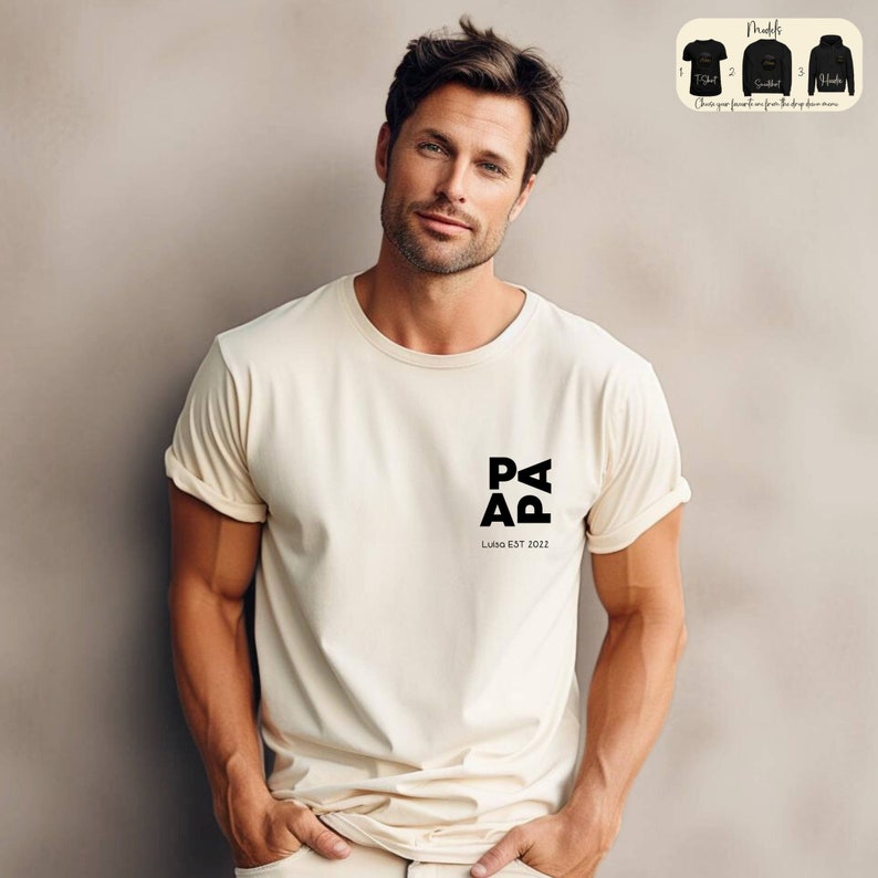Op de afbeelding: Een man draagt een cr&egrave;mekleurig T-shirt met een zwart logo met de tekst "PAPA LUKAS EST 2012".