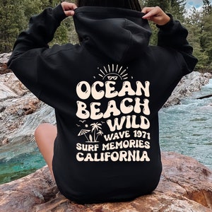 May include: Black hooded sweatshirt with a white graphic print that says "Ocean Beach Wild Wave 1971 Surf Memories California".