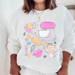 May include: White sweatshirt with a graphic design of a pink and purple stand mixer, a rolling pin, a whisk, a donut, a macaron, a cake, and the words "Love Baking Mama".