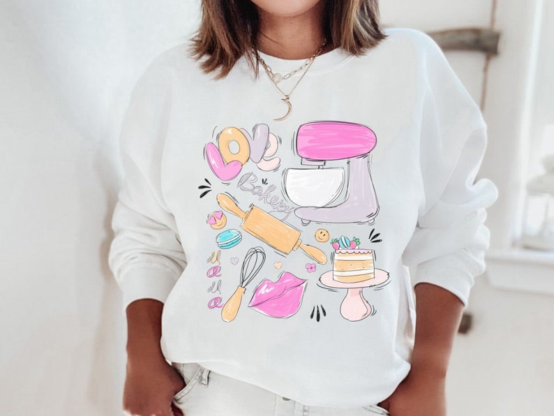 May include: White sweatshirt with a graphic design of a pink and purple stand mixer, a rolling pin, a whisk, a donut, a macaron, a cake, and the words "Love Baking Mama".
