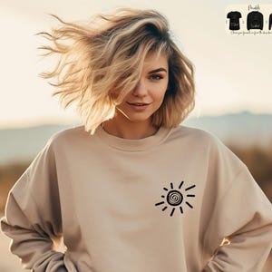 Op de afbeelding: Een beige sweatshirt met een zwarte zon-afbeelding op de voorkant. De zon is gestileerd met een spiraal in het midden en stralen die naar buiten uitstrekken.