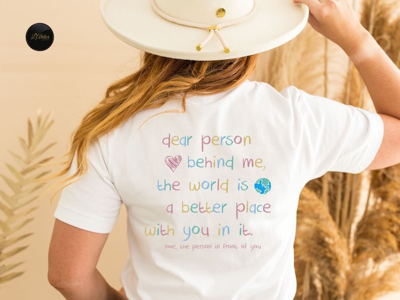 May include: White t-shirt with a colorful message that reads "dear person behind me, the world is a better place with you in it. love, the person in front of you".