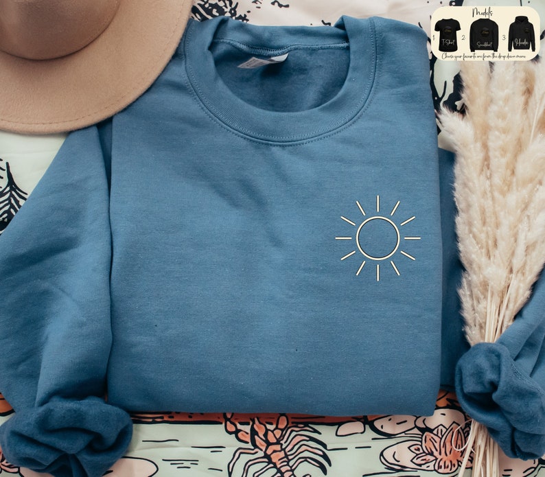 May include: A blue sweatshirt with a white sun graphic on the front. The sweatshirt is folded and laid on a patterned surface.