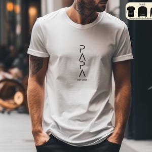 May include: White t-shirt with black text that reads "PAPA EST 2025" with the letters arranged vertically in a geometric pattern.