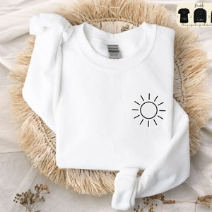 May include: White sweatshirt with a simple black sun graphic on the front. The sweatshirt is made of a soft, comfortable material.