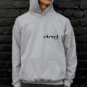 May include: A gray hooded sweatshirt with the word "dad" printed on the front in black. The text is stylized with a curved font and the words "est 2022" are printed below.