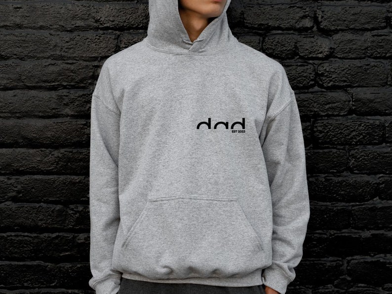 May include: A gray hooded sweatshirt with the word "dad" printed on the front in black. The text is stylized with a curved font and the words "est 2022" are printed below.