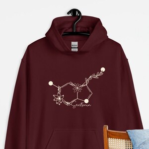 May include: Burgundy hoodie featuring a white floral serotonin molecule graphic. The graphic is a stylised chemical structure of serotonin with flowers and leaves.