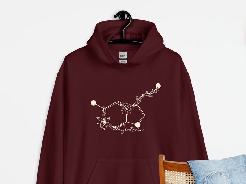 May include: Burgundy hoodie featuring a white floral serotonin molecule graphic. The graphic is a stylised chemical structure of serotonin with flowers and leaves.