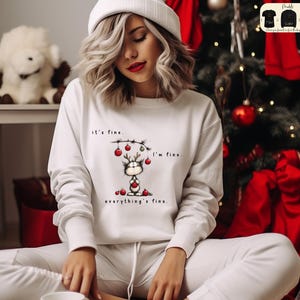 May include: White sweatshirt with a Christmas-themed graphic. The graphic features a cat with reindeer antlers, holding a heart, and surrounded by ornaments. Text on the sweatshirt reads "it's fine, I'm fine, everything's fine."