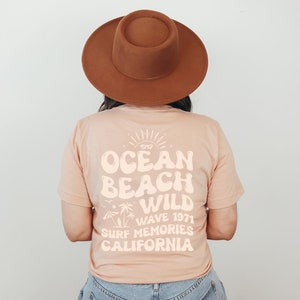 May include: A peach-colored t-shirt with a white graphic design that says "Ocean Beach Wild Wave 1971 Surf Memories California". The graphic design features a sun, palm trees, and a surfboard.