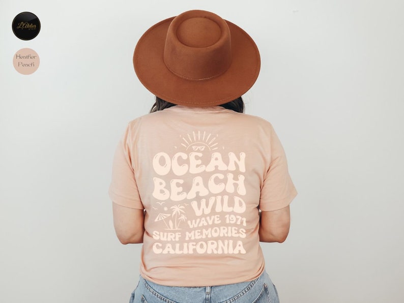 May include: A peach-colored t-shirt with a white graphic design that says "Ocean Beach Wild Wave 1971 Surf Memories California". The graphic design features a sun, palm trees, and a surfboard.