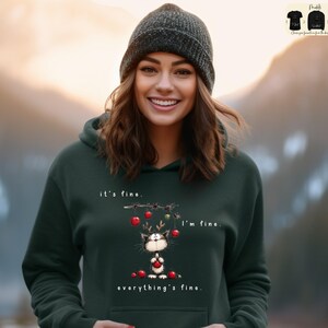 May include: Dark green hoodie with a graphic of a cat wearing reindeer antlers and holding a red ornament. The text on the hoodie reads "it's fine, I'm fine, everything's fine."