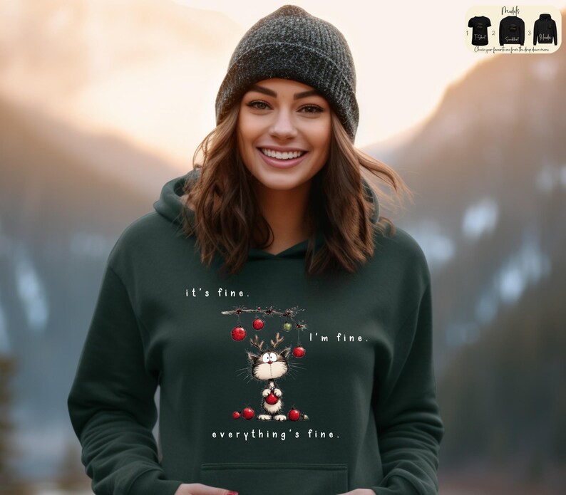 May include: Dark green hoodie with a graphic of a cat wearing reindeer antlers and holding a red ornament. The text on the hoodie reads "it's fine, I'm fine, everything's fine."