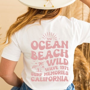 May include: White t-shirt with a pink graphic design that says "Ocean Beach Wild Wave 1971 Surf Memories California".