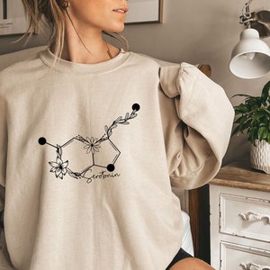 May include: A beige sweatshirt featuring a black graphic of a serotonin molecule with flowers and leaves. The text "Serotonin" is printed below the molecule.