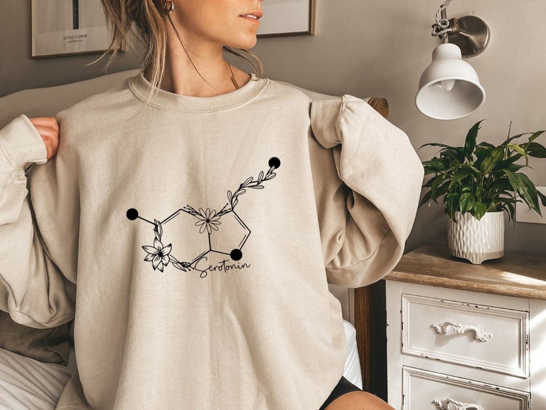 May include: A beige sweatshirt featuring a black graphic of a serotonin molecule with flowers and leaves. The text "Serotonin" is printed below the molecule.