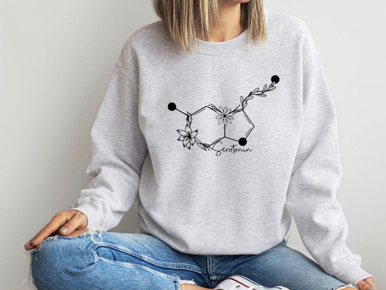 May include: A grey sweatshirt featuring a black floral design of the chemical structure of serotonin. The design includes the word "Serotonin" in black text.