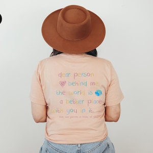 May include: A peach-colored t-shirt with a rainbow-colored message that reads "dear person behind me the world is a better place with you in it. love, the person in front of you".