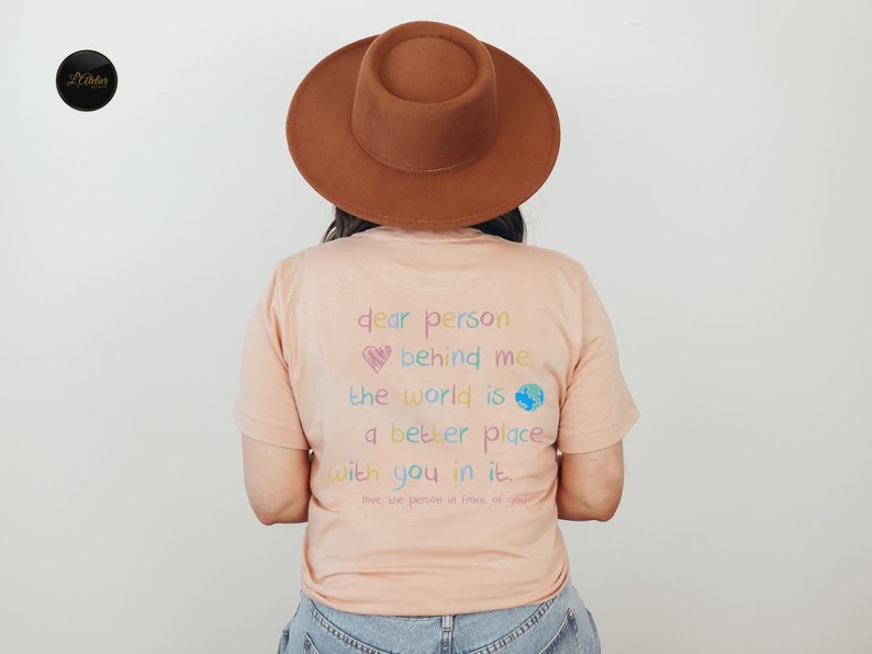 May include: A peach-colored t-shirt with a rainbow-colored message that reads "dear person behind me the world is a better place with you in it. love, the person in front of you".