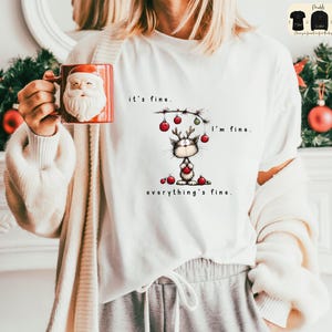 May include: White t-shirt with a cartoon cat wearing reindeer antlers and holding a red ornament. The text on the shirt reads "it's fine. I'm fine. everything's fine." A red Santa mug is held in the hand.