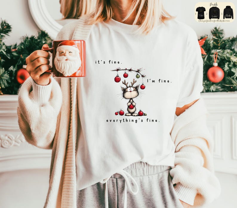 May include: White t-shirt with a cartoon cat wearing reindeer antlers and holding a red ornament. The text on the shirt reads "it's fine. I'm fine. everything's fine." A red Santa mug is held in the hand.