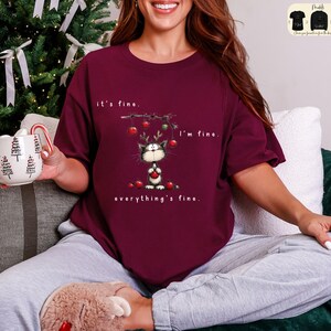 May include: Burgundy t-shirt with a whimsical cat graphic wearing reindeer antlers and holding a red ornament. The text reads "it's fine, I'm fine, everything's fine." A Christmas-themed design.
