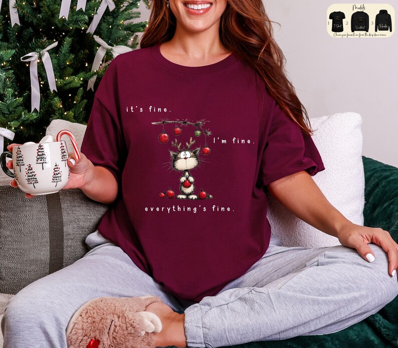 May include: Burgundy t-shirt with a whimsical cat graphic wearing reindeer antlers and holding a red ornament. The text reads "it's fine, I'm fine, everything's fine." A Christmas-themed design.