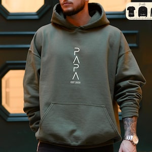 May include: A green hooded sweatshirt with the word "PAPA" written vertically in white, with the letters forming triangles. The text "EST 2025" is written below the word "PAPA".