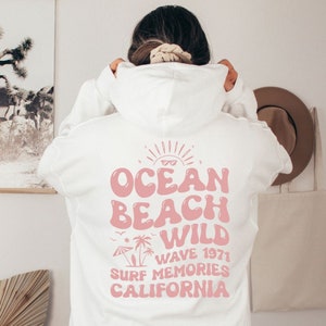 May include: White hoodie with a pink graphic print on the back that says "Ocean Beach Wild Wave 1971 Surf Memories California".