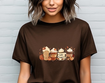 Puede incluir: Una camiseta marrón con un gráfico de tres tazas de café de temática otoñal con crema batida y chispas. Las tazas tienen el texto "Hello Pumpkin" en una de ellas. El gráfico también incluye calabazas y hojas.