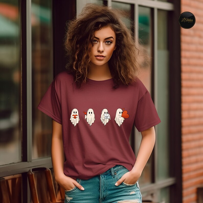 May include: A maroon t-shirt with a graphic of four white ghosts. The ghosts are in a row and each has a different expression. The first ghost is blowing a kiss, the second ghost is smiling, the third ghost is holding a heart, and the fourth ghost is holding a heart and has a surprised expression.