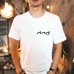 May include: White t-shirt with a black graphic that says "dad est. 2023".