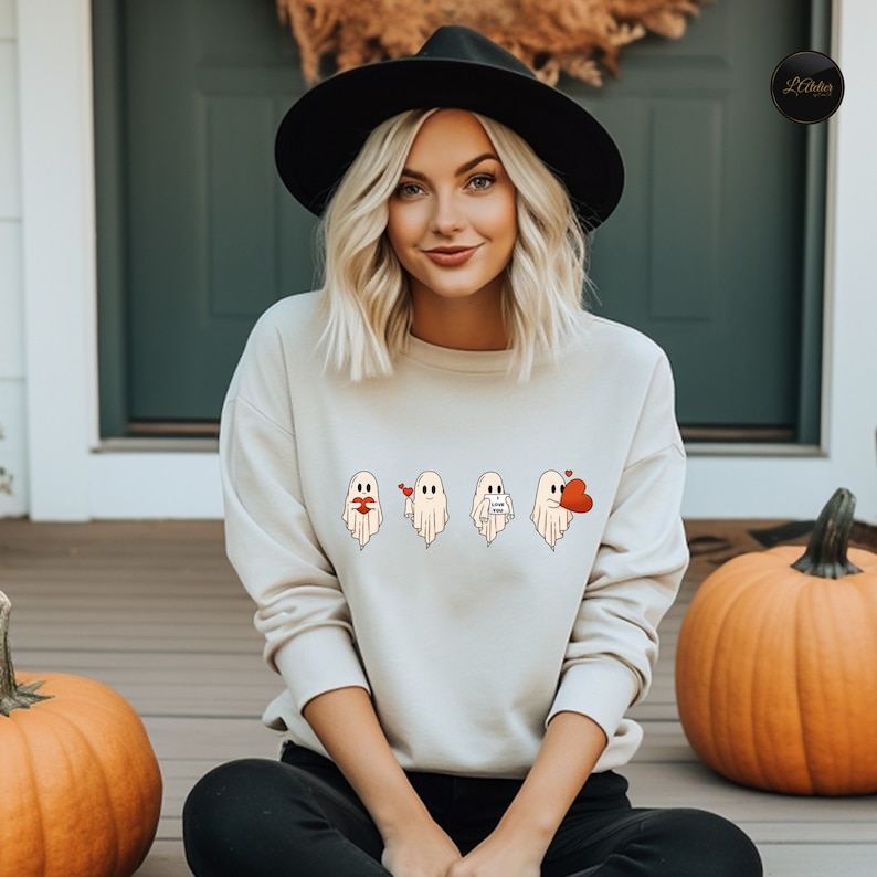 May include: A light beige sweatshirt with a graphic of four ghosts. The ghosts are wearing different expressions, including a heart, a kiss, a love note, and a balloon. The sweatshirt is being worn by a person with blonde hair and a black hat.