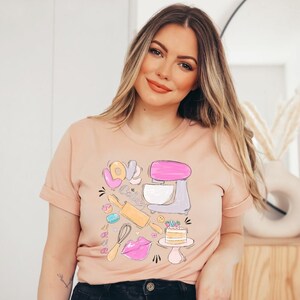 May include: A light pink t-shirt with a graphic design of a pink and gray stand mixer, a rolling pin, a whisk, a donut, a macaron, a cake, and the word "LOVE" in pink, yellow, and blue letters.