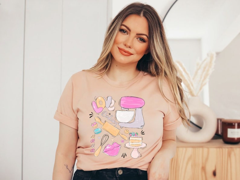 May include: A light pink t-shirt with a graphic design of a pink and gray stand mixer, a rolling pin, a whisk, a donut, a macaron, a cake, and the word "LOVE" in pink, yellow, and blue letters.