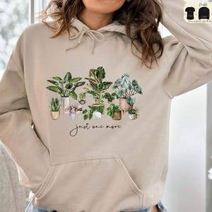 Peut inclure: Sweat à capuche beige avec une illustration à l'aquarelle de plantes en pot. Le motif comprend diverses plantes vertes dans différents pots, avec le texte manuscrit "just one more" en dessous. Le sweat à capuche a une poche avant et une capuche à cordon.