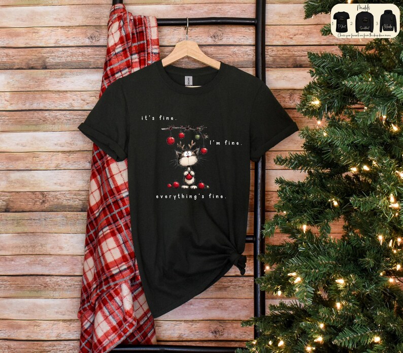 May include: Black t-shirt with a Christmas cat graphic. The cat is wearing reindeer antlers and holding ornaments. Text on the shirt reads "it's fine. I'm fine. everything's fine."